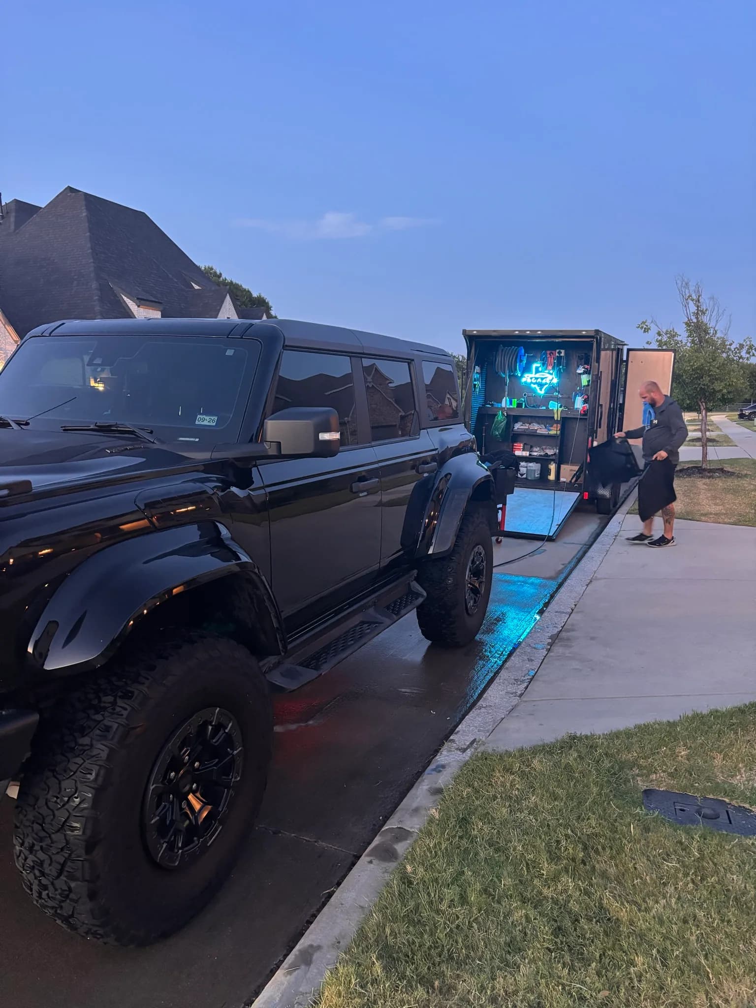 Black Ford Bronco Raptor beside the lit mobile detailing trailer at dusk.