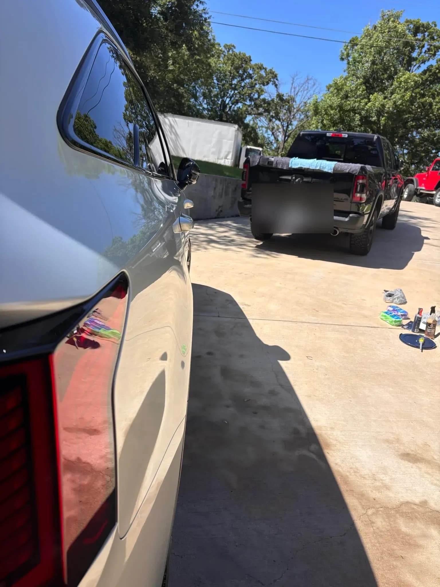 Polished rear quarter panel of a white Kia SUV reflecting the driveway, with a black Ram pickup being detailed in the background.