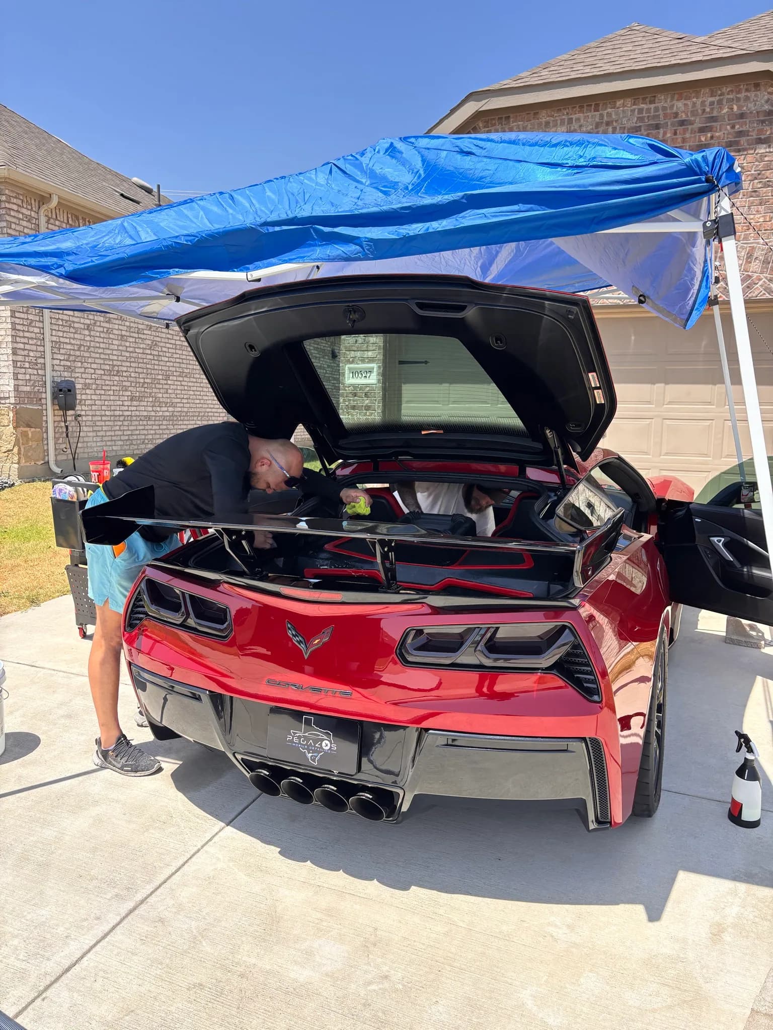 Detailer working at the open rear hatch of a red Corvette under a canopy.