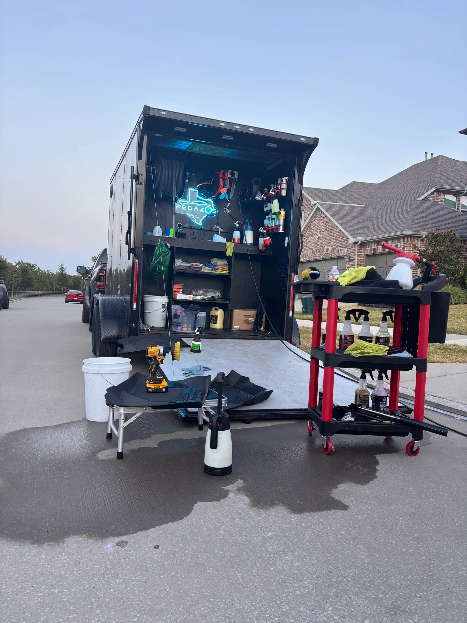 The mobile detailing trailer set up in a driveway at dusk with carts and tools laid out.